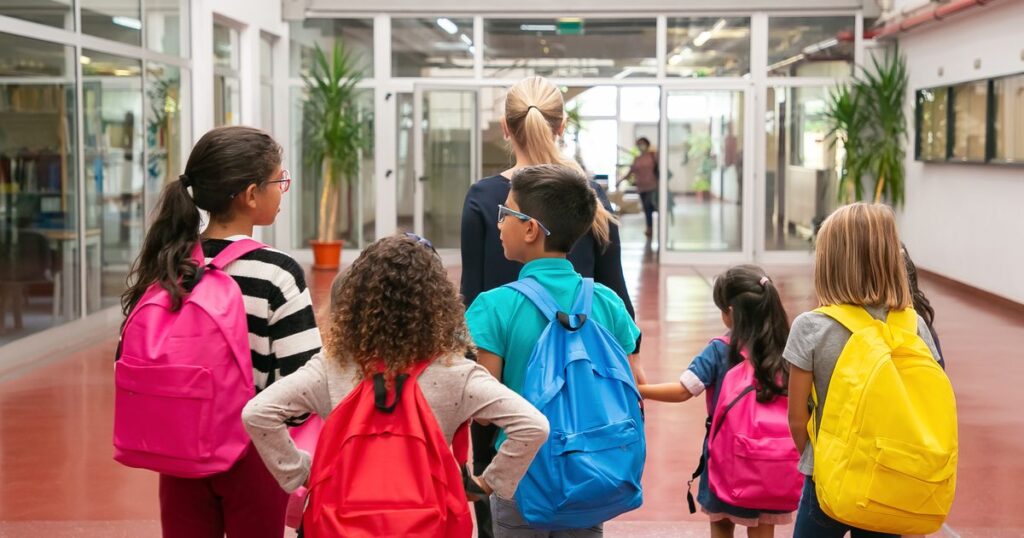 Alunos com mochilas caminhando no corredor da escola acompanhados por uma professora
